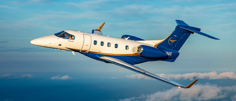 A Phenom 300 in flight with sunset kissed light clouds below. a golden hour reflects off the plane highlighting the gold accent that cuts down the side of the plane between the white painted body and blue tail and underbody.