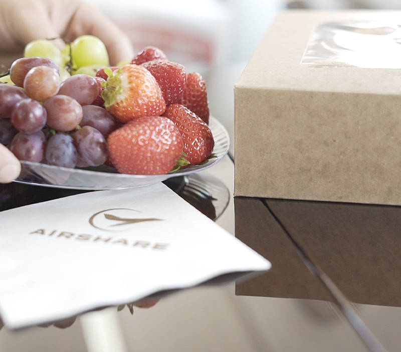 Fresh fruits and pastries in a box being placed over an Airshare napkin on plane table representing the high end flight service you get with Airshare.